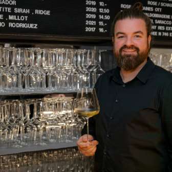Sympathischer Barkeeper der WY Weinbar mit Weinglas vor stilvollem Regal voller Gläser und Weine.