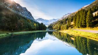 Spiegelglatter Gänglesee in Steg, umrahmt von dichten Wäldern und schneebedeckten Bergen bei Sonnenschein