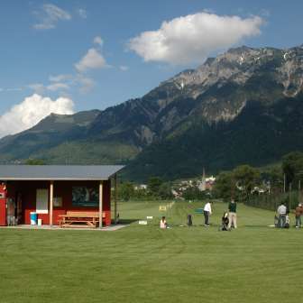 Golferinnen und Golfer auf der Driving Range in Liechtenstein mit Blick auf grüne Wiesen, ein rotes Clubhaus und die umliegende Berglandschaft bei blauem Himmel.
