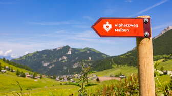 Wegweiser des Alpherzwegs mit Bergdorf Malbun im Hintergrund