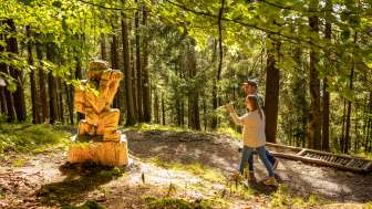 Two hikers walk past a wooden figure on the Walser Legend Trail