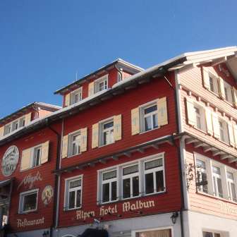 Außenansicht des Alpenhotel Vögeli Malbun mit der Elchbar im Erdgeschoss – rotes Traditionshaus mit weißer Beschriftung vor blauem Himmel.