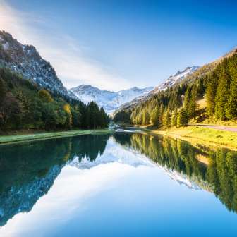 Spiegelglatter Gänglesee in Steg, umrahmt von dichten Wäldern und schneebedeckten Bergen bei Sonnenschein