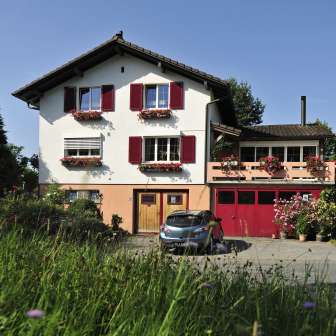 Charmante Außenansicht des Bed & Breakfast Büchel mit roten Fensterläden und Blumenwiese im Vordergrund.