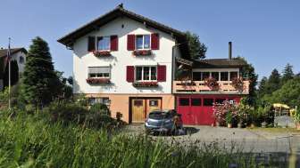 Charmante Außenansicht des Bed & Breakfast Büchel mit roten Fensterläden und Blumenwiese im Vordergrund.