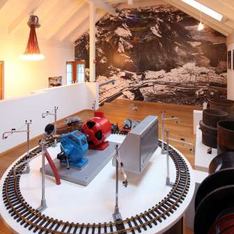 Modell einer Turbine auf einer Ausstellungsplattform im Lawena Museum, im Hintergrund historische Schwarzweißaufnahmen.