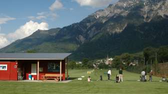 Golferinnen und Golfer auf der Driving Range in Liechtenstein mit Blick auf grüne Wiesen, ein rotes Clubhaus und die umliegende Berglandschaft bei blauem Himmel.
