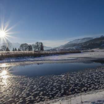 Sonnenaufgang am Egelsee in Mauren im Winter mit glitzernder Eisfläche, Schnee und sonniger Bergkulisse.