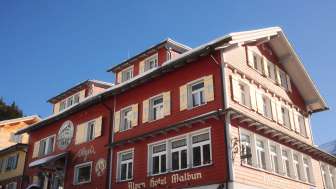 Außenansicht des Alpenhotel Vögeli Malbun mit der Elchbar im Erdgeschoss – rotes Traditionshaus mit weißer Beschriftung vor blauem Himmel.