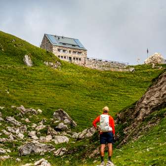 Wanderer mit rotem Rucksack blickt auf die Pfälzerhütte inmitten grüner Berglandschaft